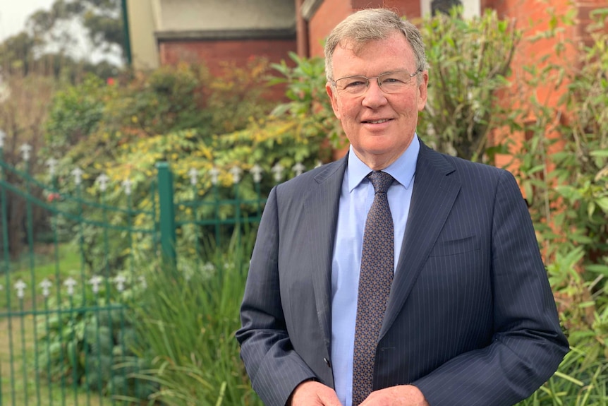 Bruce Mann stands in front of a red-brick building and garden.