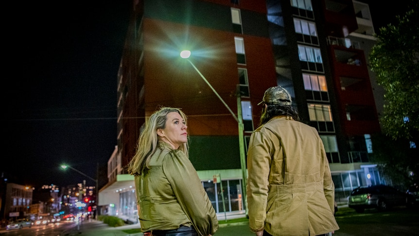 Under a bright streetlight, a woman looks away from a modern apartment block as a man looks towards it