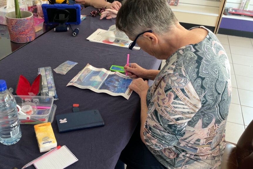 A woman looks down at craft at a table. She puts coloured gems in a dolphin shape. She has short grey hair and glasses