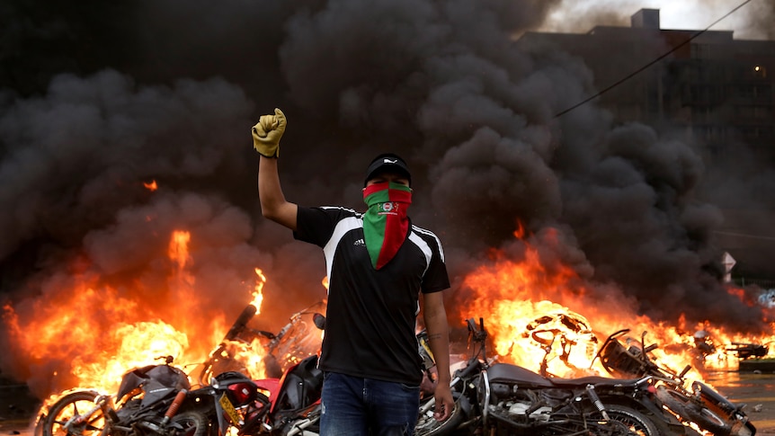 A man walks holding his fist up in front of burning motorbikes and wreckage