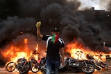 A man walks holding his fist up in front of burning motorbikes and wreckage