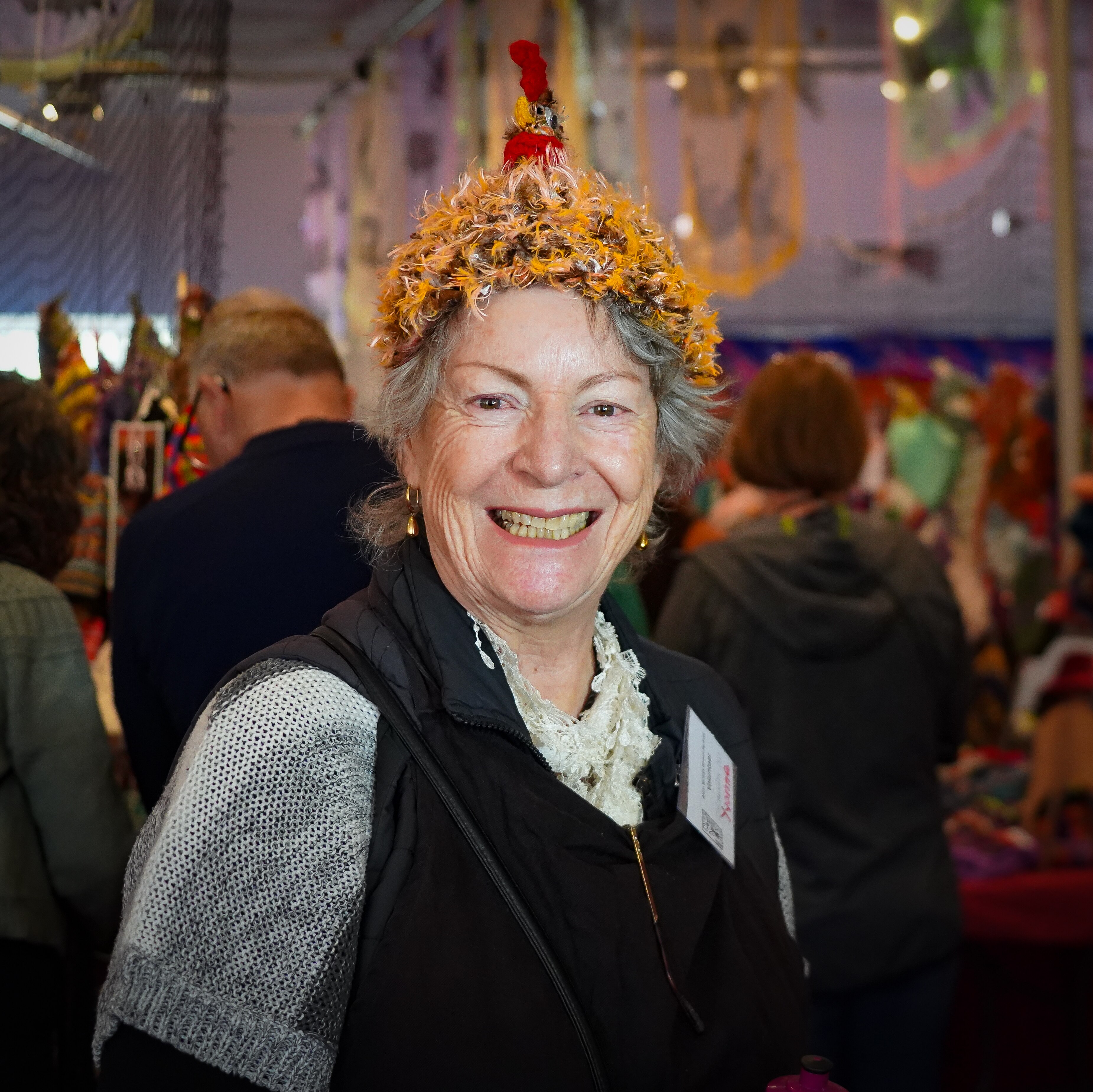 A woman smiles at the camera while wearing an orange and red beanie, it has been knitted to look like a roosting chicken.