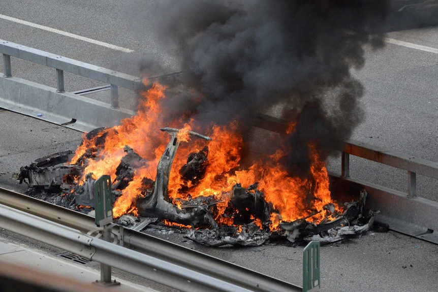 A fire blazes on an empty road, with black smoke billowing above a mangled wreck.