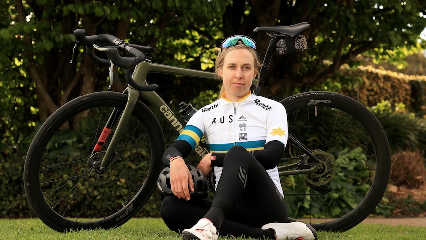 A slightly smiling blonde woman, hair tied back, aviators on top of head, sitting in front of bike, wears white cycling jersey.