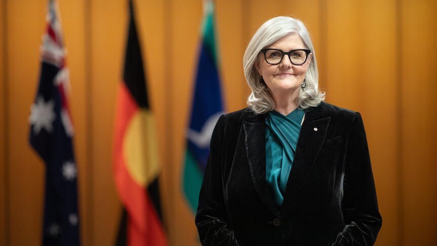 Sam Mostyn stands in front of the Australian, Aboriginal and Torres Strait Islander flags