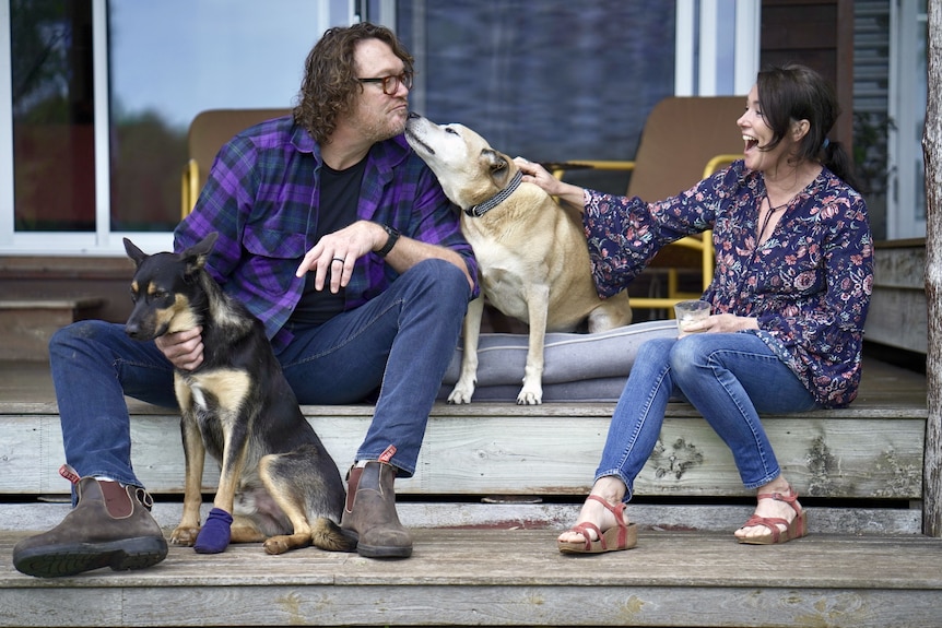 A man wearing glasses leans in for a 'kiss' from his dog as a woman laughs. The pair sit on a deck