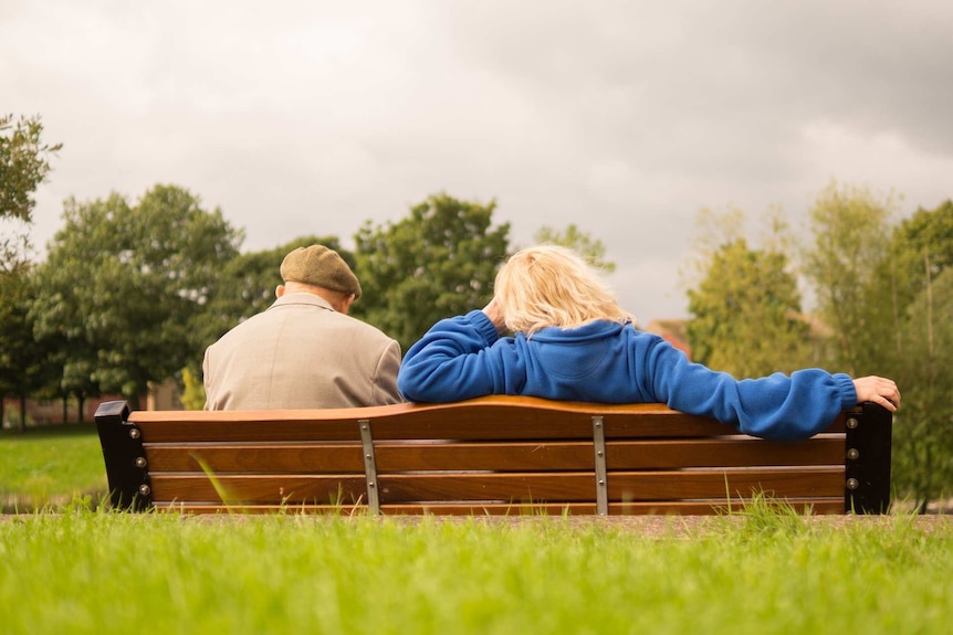 Two people seen from behind sit on a park bench, with trees and grass surrounding them.