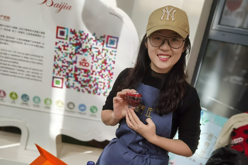 A smiling woman wearing a gold baseball cap shows a product from her shop as she poses for a photograph.