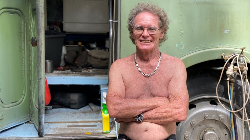 Shirtless man sitting outside an open bus door with his arms crossed.