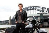 Austin Butler in a leather jacket stands by a motorbike, with the Sydney Harbor Bridge behind him