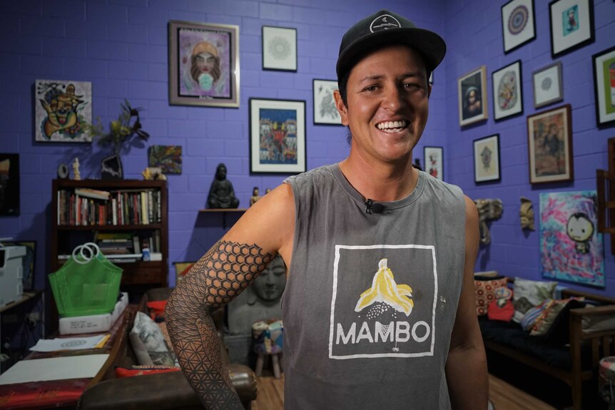 Rhys Shearer smiles at the camera. He has a tattoo on his arm, and behind him are many framed drawings.