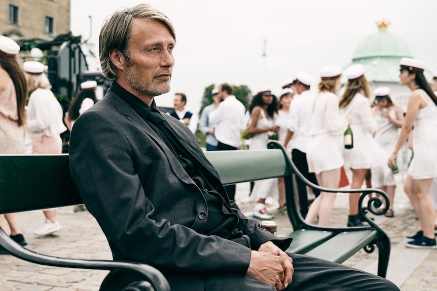Pensive-looking Mads Mikkelsen wearing dark suit and shirt and sitting on bench, with crowd of young adults in background.