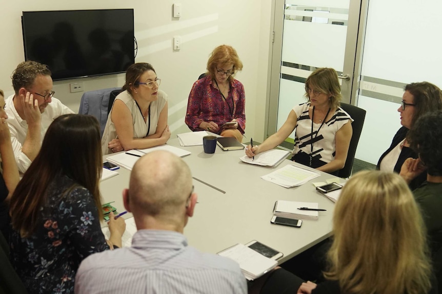 Ten people sitting at desk with notepads and phones.