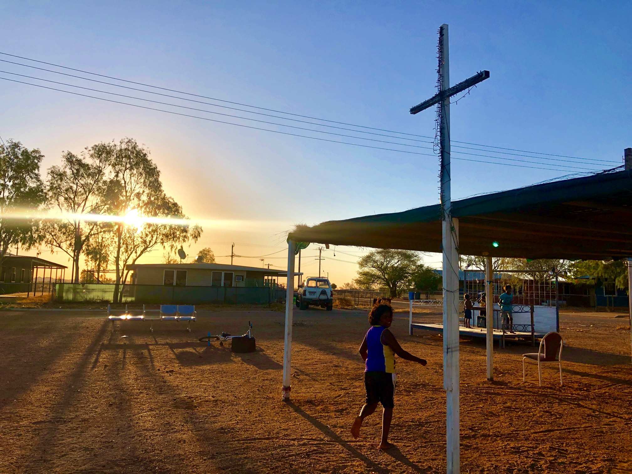 A young Aboriginal boy runs beneath a cross in a remote community. A stage is in the background with children playing on it.