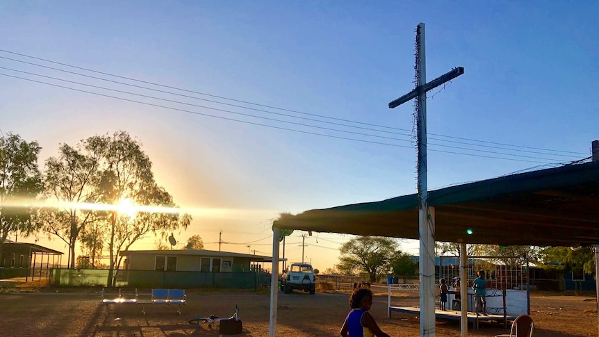 A young Aboriginal boy runs beneath a cross in a remote community. A stage is in the background with children playing on it.