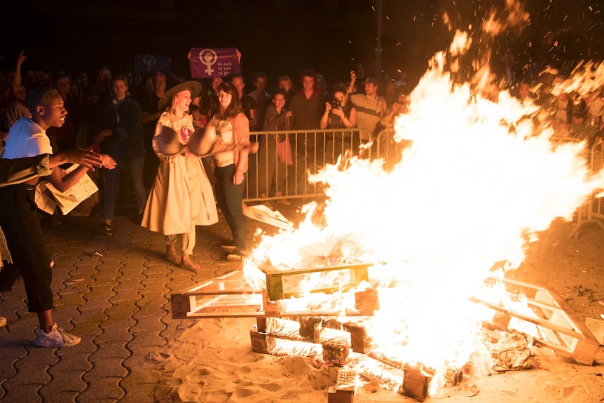 A bra is thrown into a bonfire, burning on a paved area while a crowd watches on.