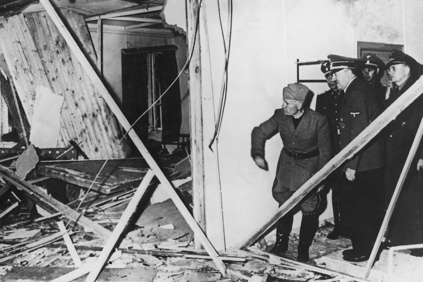 Black and white photo of five men crowded together looking into room with crumbling roof, fallen wires and debris over floor.