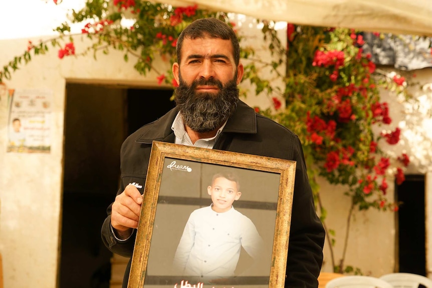 A man with dark hair and a beard holds a photo of a young boy in a white shirt outside under a shade.