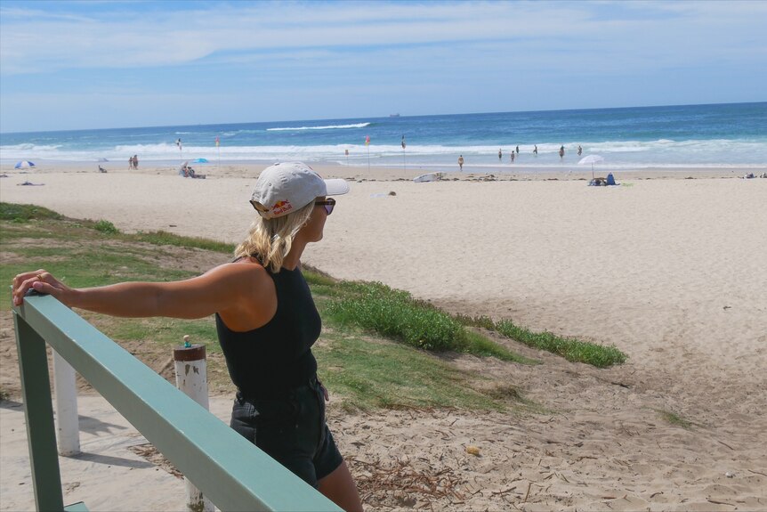A woman wearing a cap and sunglasses holding a railing looking at a beach