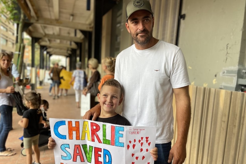 Man in cap and white t-shirt standing next to child holding sign