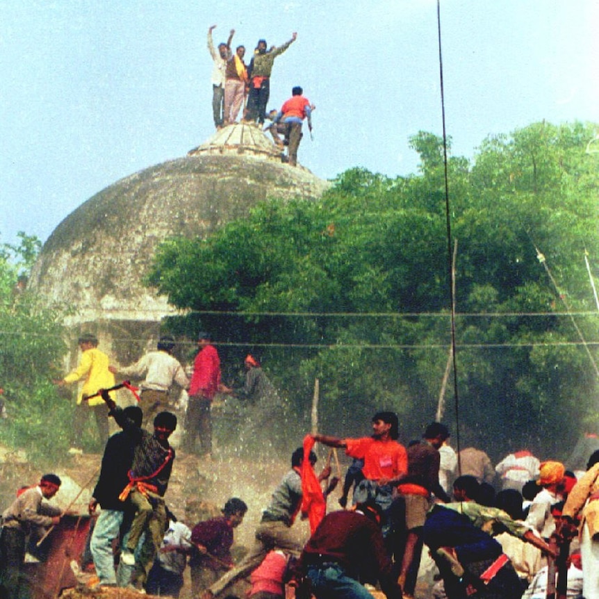 People stand on the top dome of a mosque while others climb over walls in the foreground