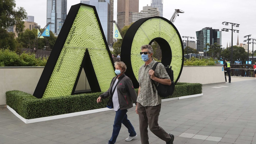 A man and a woman wearing masks walk past a large "AO" sign at the Australian Open.