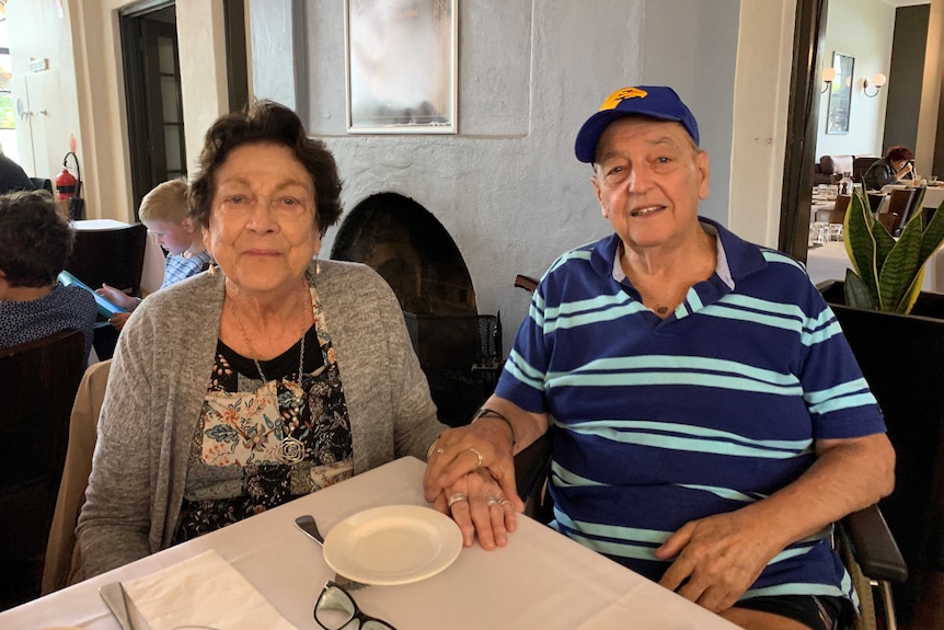 An older couple sit smiling at a table, holding hands.
