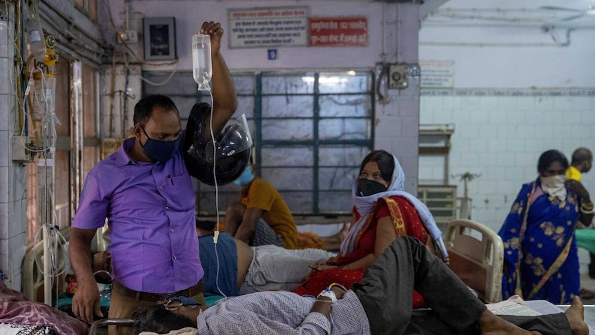 A man holds up an intravenous (IV) drip being used to treat a man lying on a bed as a woman sitting on a hospital bed watches on