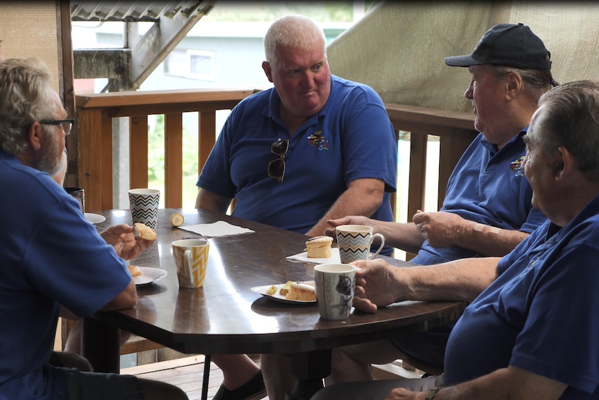 A group of older men sit at tables having cups of coffee and chatting.