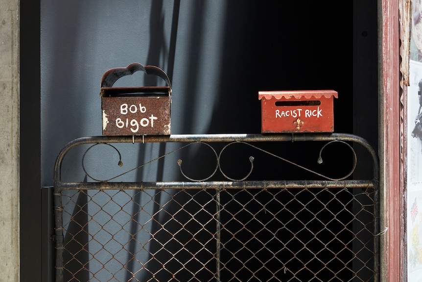 Two metal letterboxes, one dark, one red, sit on top of an iron gate, one reading "Bob Bigot", the other "Racist Rick"