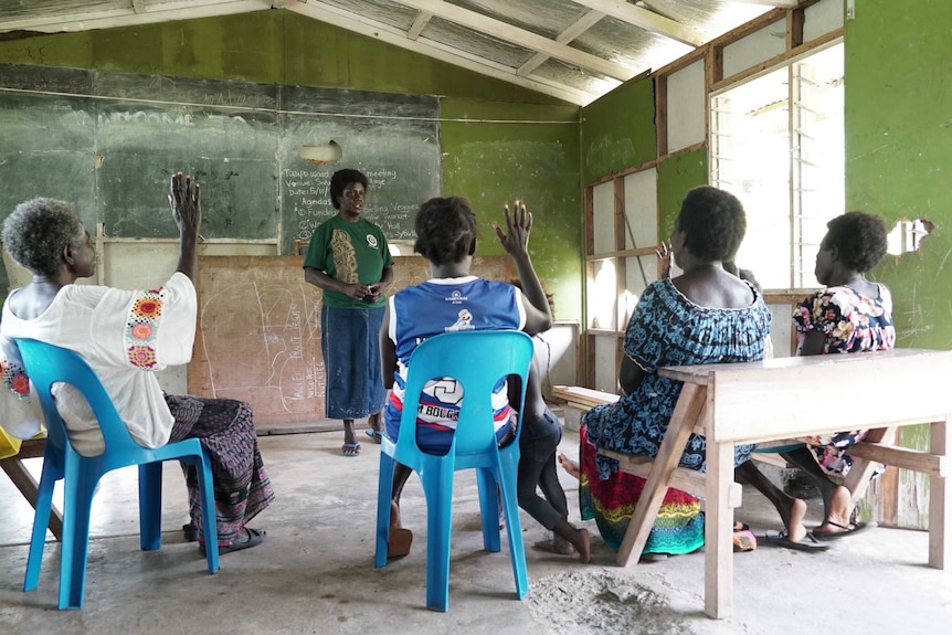 Doreen Nauvana stands infront of a blackboard watching four other women sitting with their hands raised in the air.