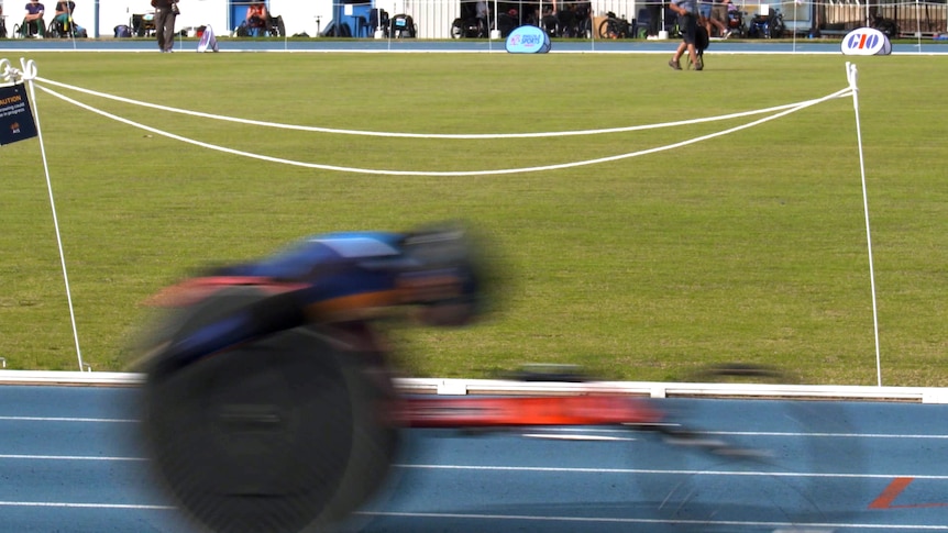 A racing wheelchair speeds past the camera along a racing track. It is daytime. The rider's speed reduces them to a blur.