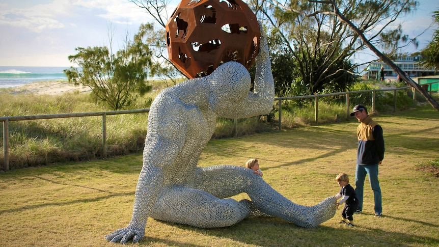 Father with two toddlers looking at chain mesh statue of man holding up steel globe.