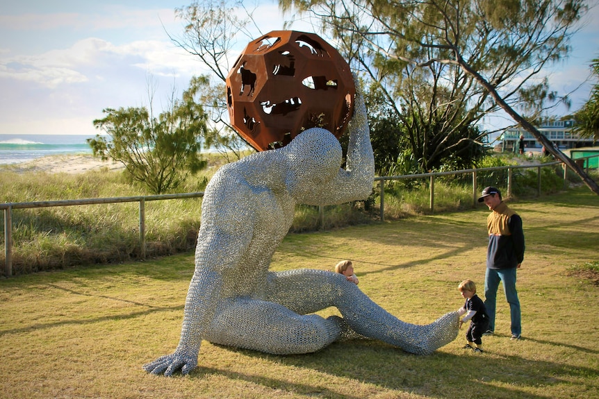 Father with two toddlers looking at chain mesh statue of man holding up steel globe.