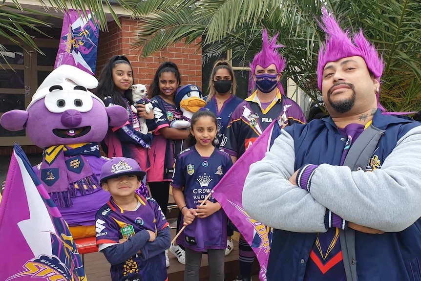 A family of seven dressed in Melbourne Storm gear pose with a purple Smurf doll in front of a house.
