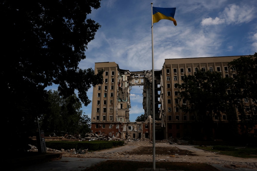 View of destroyed building with Ukrainian flag flying on pole in front.