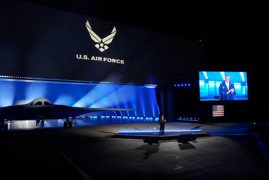 a stealth bomber on a stage with a US air force logo above it and a person speaking at centre stage