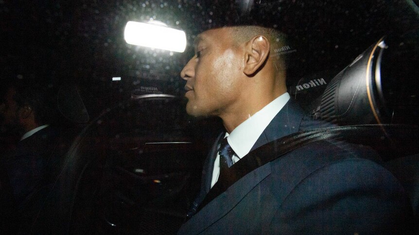 A man in a suit sits in the back seat of a car leaving a building car park.