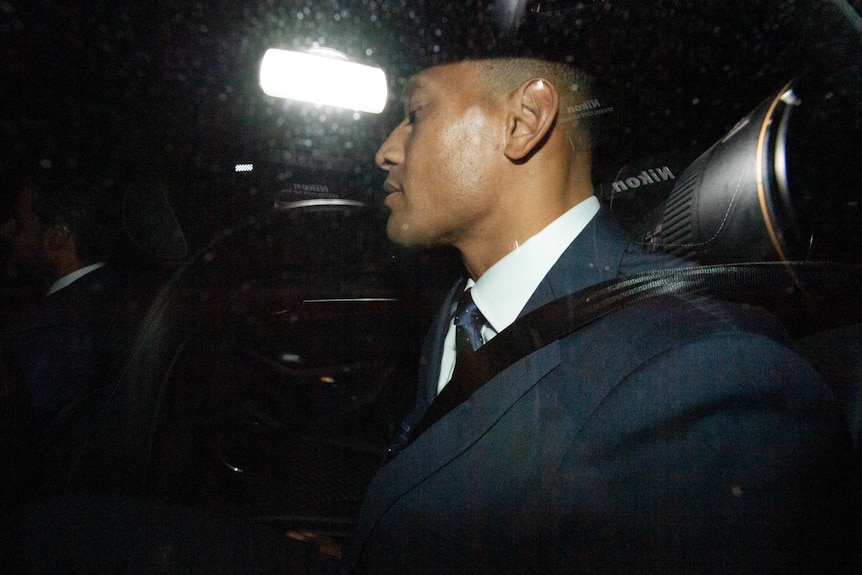 A man in a suit sits in the back seat of a car leaving a building car park.