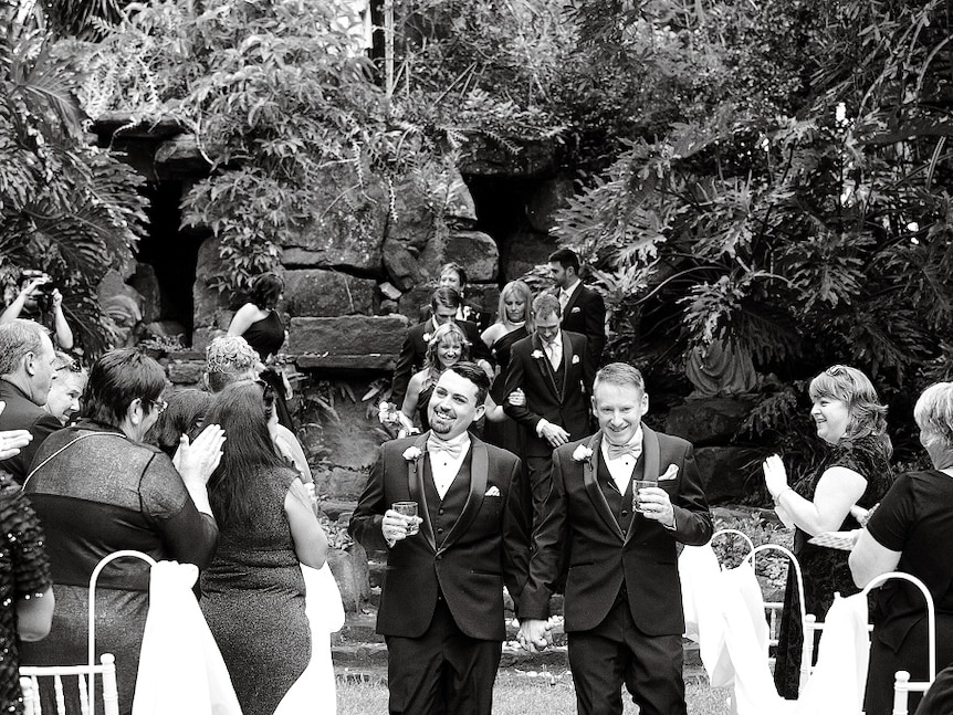 Two men leaving a commitment ceremony smiling at loved ones and holding hands