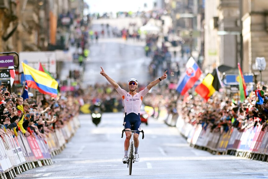 A cyclist raises his arms in triumph as he rolls across the line with crowds cheering at the end of a race.
