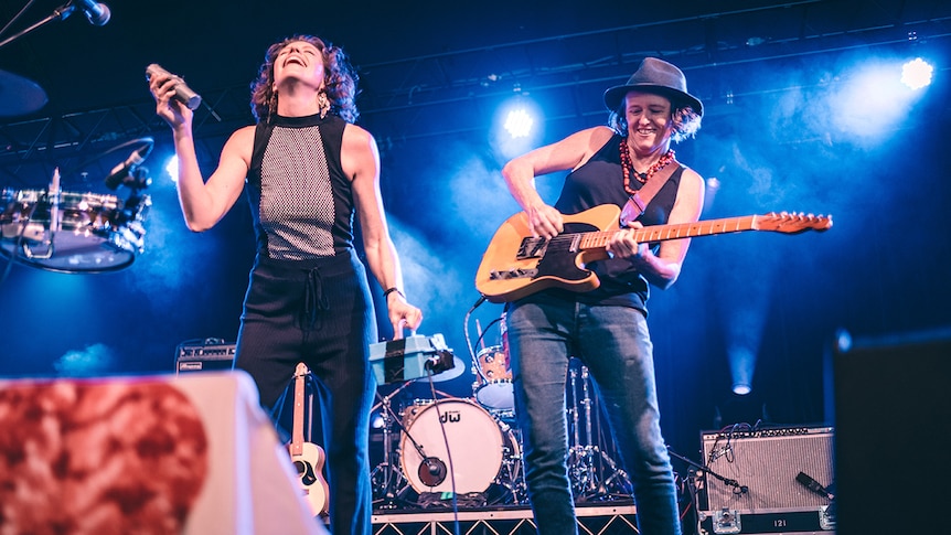 Two women of the band Hussy Hicks grin while playing on stage. One holds hand percussion, another plays guitar