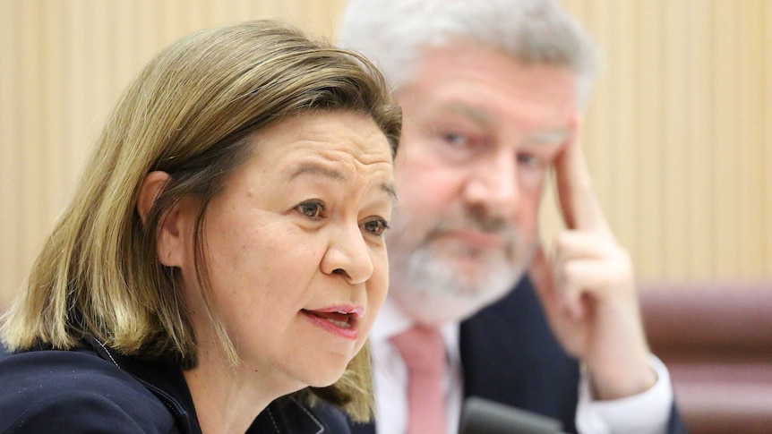 A close-up photo of Guthrie speaking, while Mitch Fifield looks her way in the background.