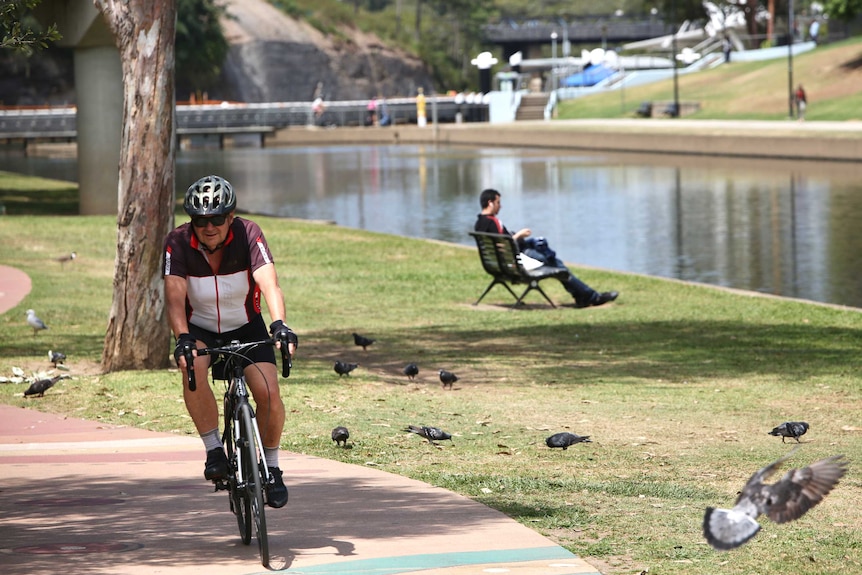 A cyclist rounds the bend on a pathway next a river, a man sits on a bench behind him.