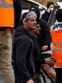 A close-up of a man wearing dark clothes and a plastic goggle stands with people wearing high-viz jackets.