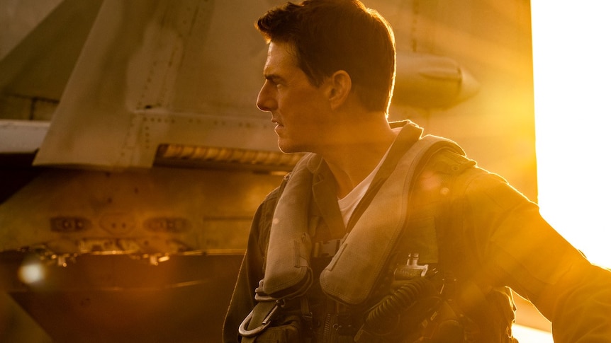 Man wearing military pilot uniform standing next to grey plane with orange sunlight streaming over his shoulder