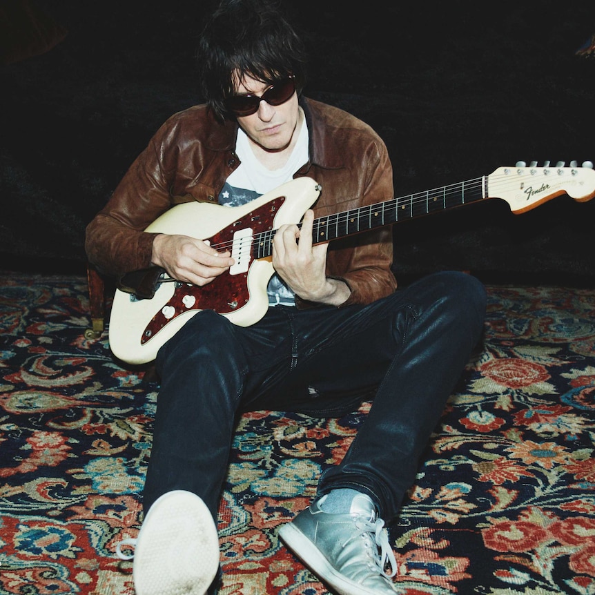 Jason Pierce is photographed in a brown leather jacket with sunglasses on playing his electric guitar