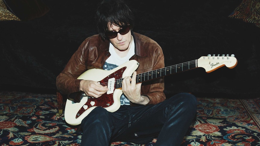 Jason Pierce is photographed in a brown leather jacket with sunglasses on playing his electric guitar