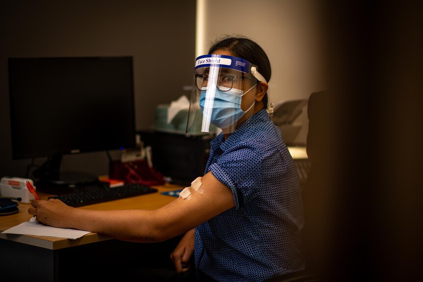 A woman wearing a face shield and mask with bandages up her arms looks at the camera.