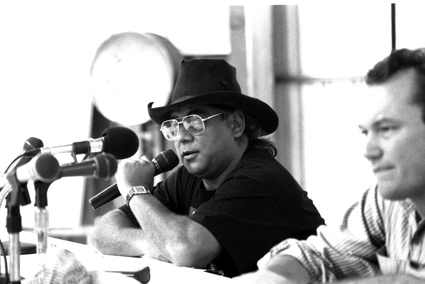 A black and white photo of Tilmouth wearing a cowboy hat, speaking into a mic at a press conference.